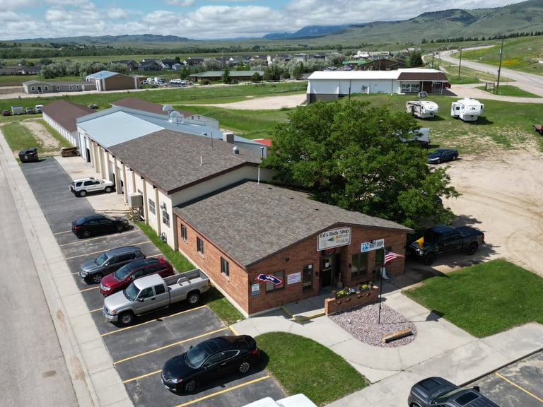 Aerial view of a commercial building complex with red brick and metal roof structures, parking lot with vehicles, surrounded by rural landscape and mountains